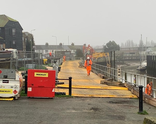 A picture of our current work at Stand Quay. A construction worker in orange hi-vis walks along a yellow path next to a river. There is construction work grey metal fences along the left hand side of the yellow path. There is a line of buildings including houses on the far left of the picture. There are two construction vehicles, one red and one yellow, at the back of the picture. The yellow one has a grabbing arm which is moving near the river. There are two boats moored on the river, one black and one white, on the right hand side of the picture. 