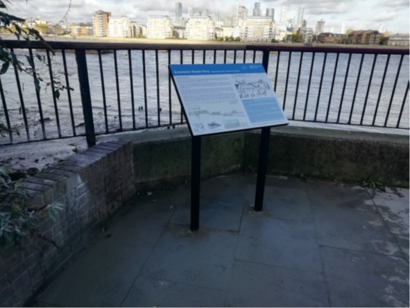 An information board on a paved walkway with railings with the River Thames behind..