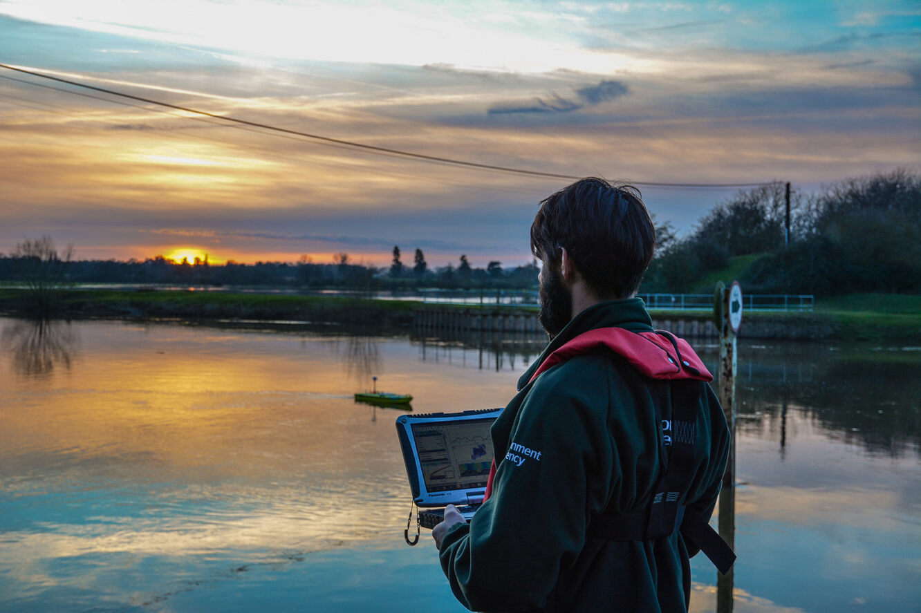 Man looking across a lake, holding a digital tablet, with an orange sunset in the horizon. 