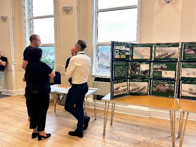 Picture 1 shows people, including Severn Trent colleagues, gathered in a group discussing the scheme, with a display board next to them with scheme designs showing. 